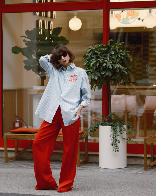 Person wearing a light blue shirt with red text and red pants standing in front of a store window.